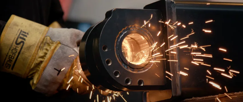 Ein Metallarbeiter mit Schutzhandschuhen schleift ein Bauteil, Funken fliegen in die Umgebung.
