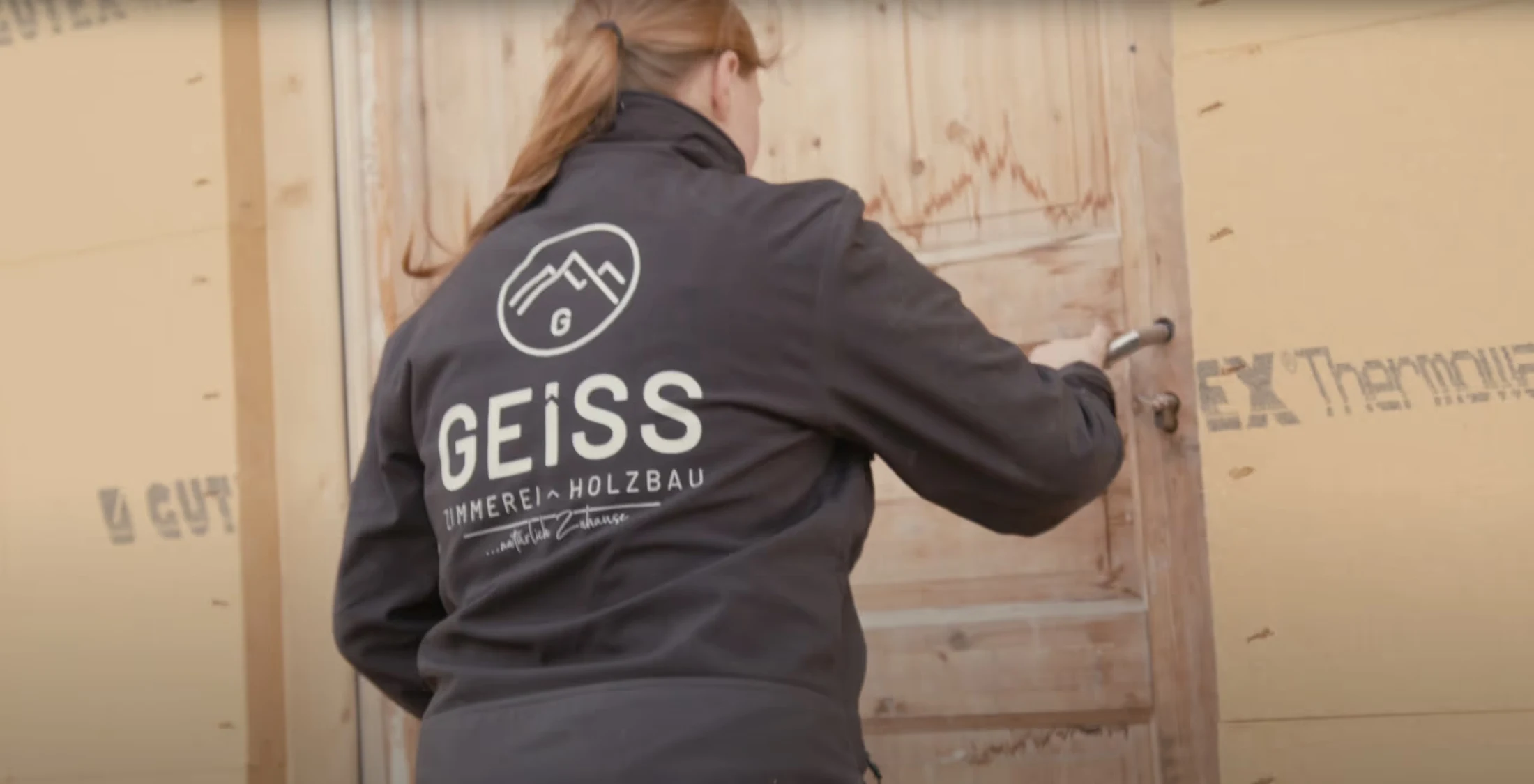 Mitarbeiterin der Zimmerei GEISS in Arbeitsjacke öffnet eine Holztür auf einer Baustelle, im Hintergrund Holzbau-Material.