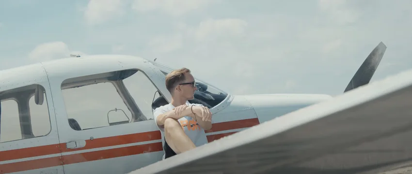 Mann mit Brille sitzt entspannt vor einem kleinen weißen Flugzeug mit roten Streifen unter blauem Himmel.