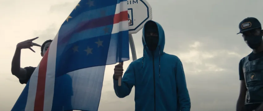 Drei Personen mit verdeckten Gesichtern, eine davon hält eine wehende Flagge von Kap Verde, vor einem Basketballkorb bei bewölktem Himmel.