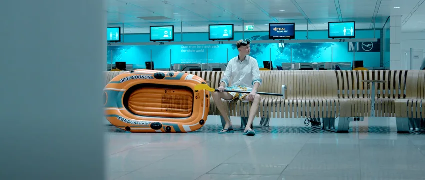 Ein Mann in Sommerkleidung sitzt mit einem orangefarbenen Schlauchboot und einem Paddel auf einer Bank in einem Flughafenterminal.