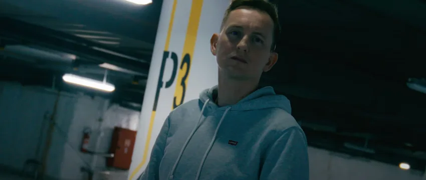 Ein Mann in grauem Hoodie steht in einer Parkgarage, umgeben von Betonwänden und markierter Säule mit der Aufschrift 'P3'.