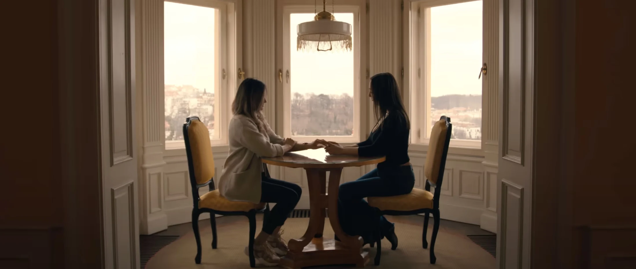 Zwei Frauen sitzen sich an einem eleganten Holztisch gegenüber, mit einem malerischen Stadtpanorama im Hintergrund.