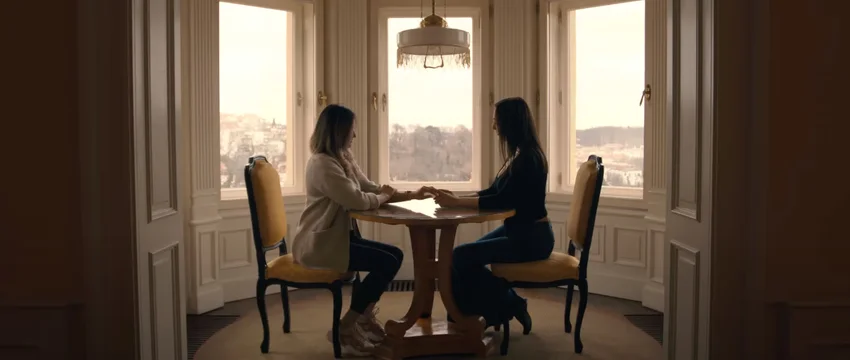 Zwei Frauen sitzen sich an einem eleganten Holztisch gegenüber, mit einem malerischen Stadtpanorama im Hintergrund.
